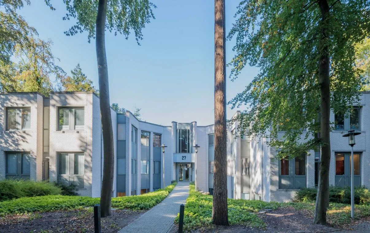 Twee lichte gebouwvleugels met daartussen een pad naar de ingang met huisnummer 27, omgeven door hoge bomen.