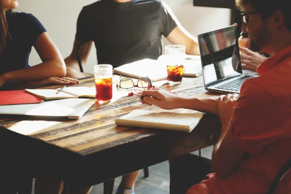 Vier mensen aan een houten tafel met laptops, notitieboeken en glazen, in gesprek over hun werk.